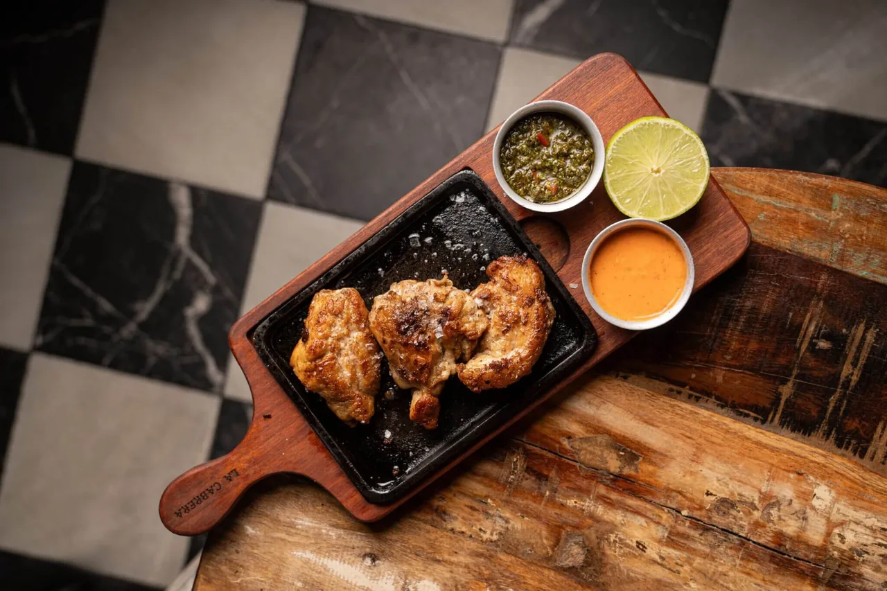 Grilled chicken on a cast-iron plate with chimichurri, a spicy cream sauce and half a lime, on a wooden paddle branded LA CABRERA over a chequered floor