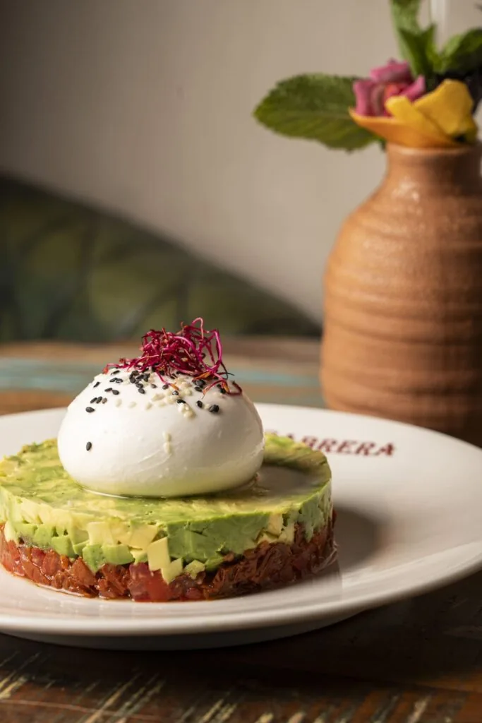 Burrata on a ring of diced avocado over tomato tartare, topped with sesame seeds and red sprouts, on a plate marked CABRERA