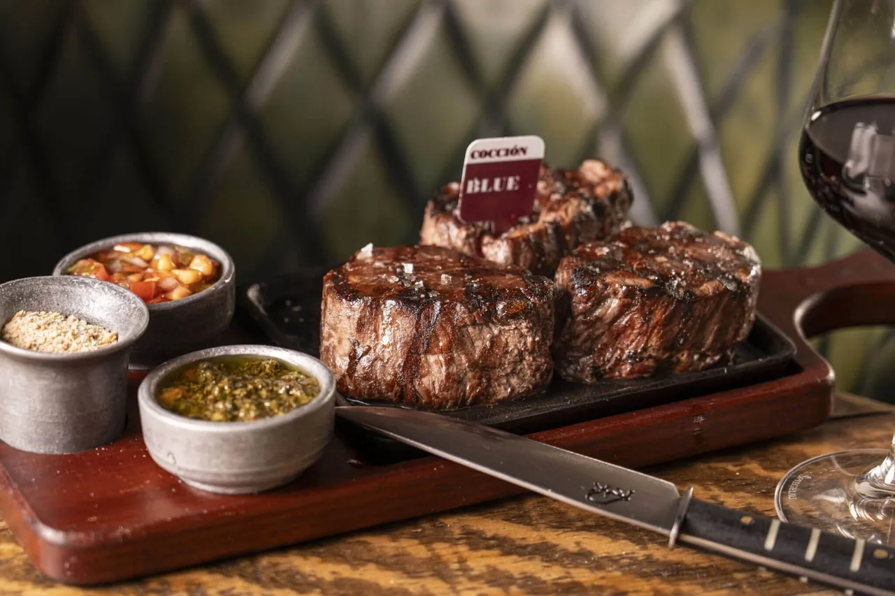 Three thick grilled steaks on a wooden board with small bowls of sauces and a steak knife