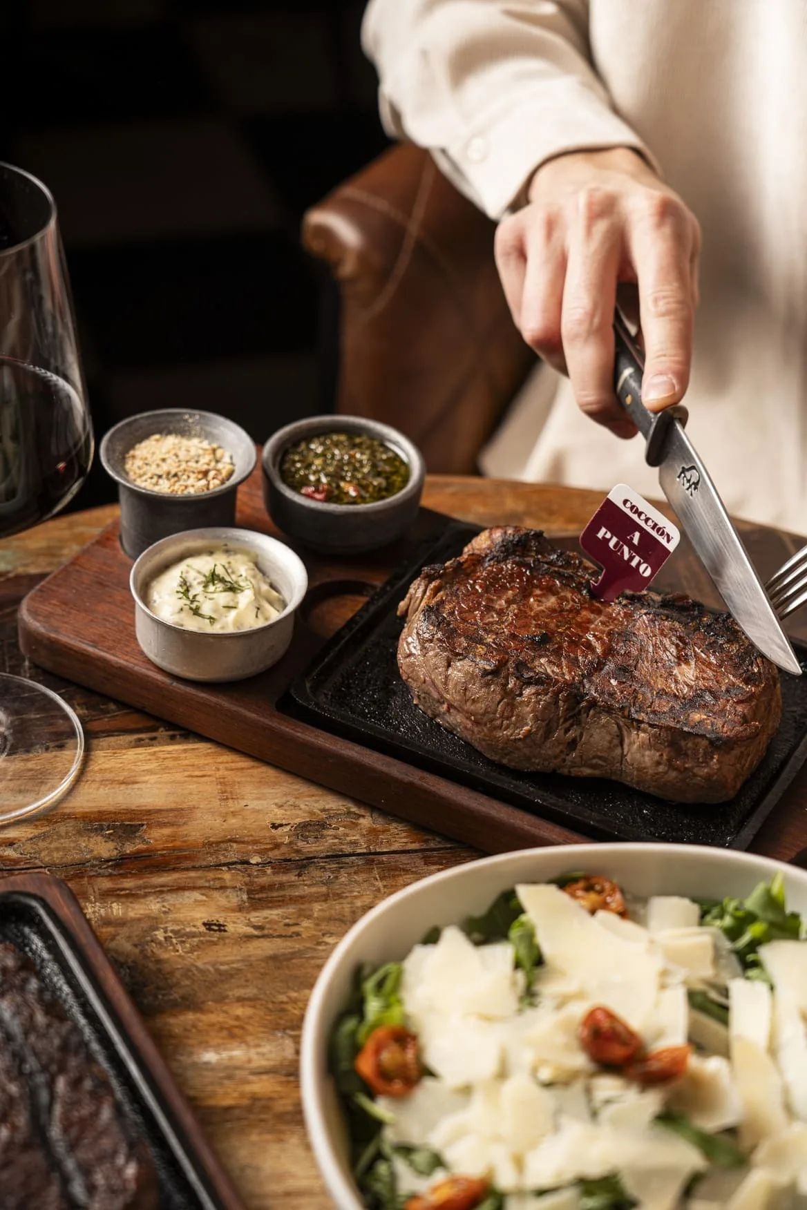A grilled steak on a cast-iron plate being cut with a knife, beside a salad