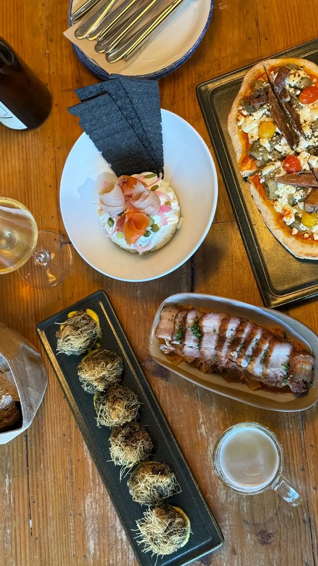 A table spread with a bowl of tartare topped with pink fish curls and black crackers, a flatbread pizza, kataifi-wrapped fried bombas, and sliced pork belly, alongside a glass of white wine and a glass of beer