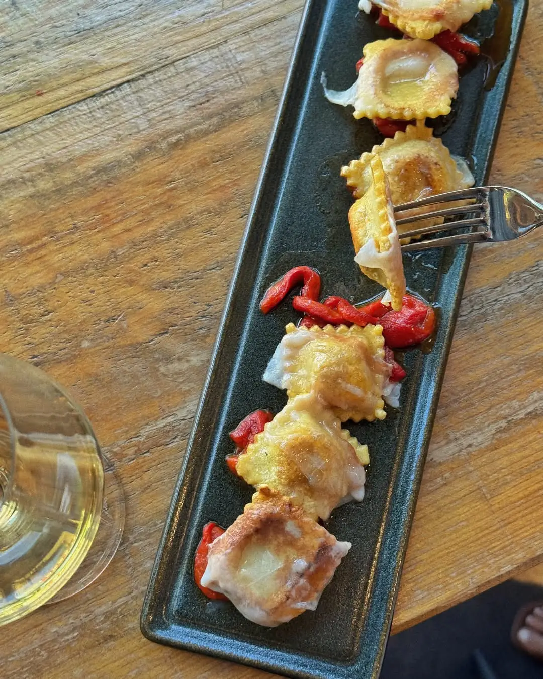 Ravioli in a light butter sauce with roasted red pepper strips on a black rectangular plate, one piece lifted on a fork