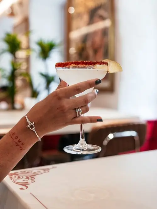 Hand holding a margarita in a coupe glass with a chili-salt rim and lime wheel, in a bright plant-filled interior