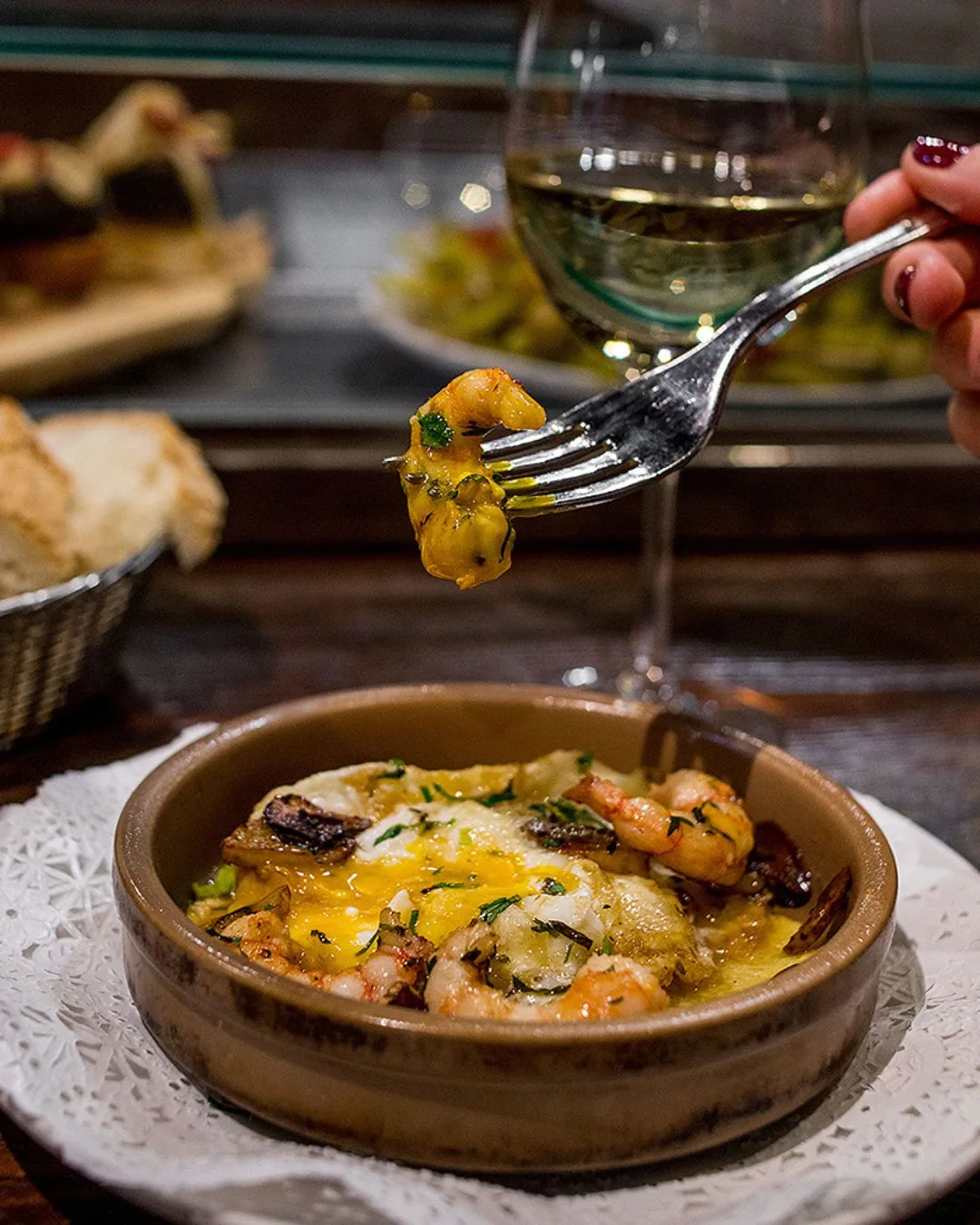 Sizzling prawns with garlic and parsley over a broken fried egg in a clay dish, with bread and white wine