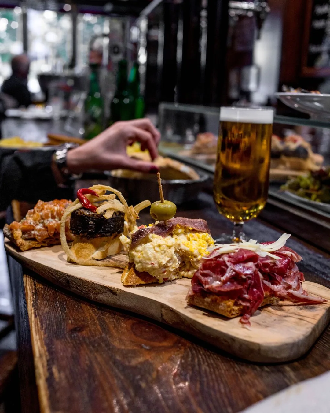 A wooden board of pinchos on bread, including cured meat, ensaladilla with anchovy and morcilla, with a beer