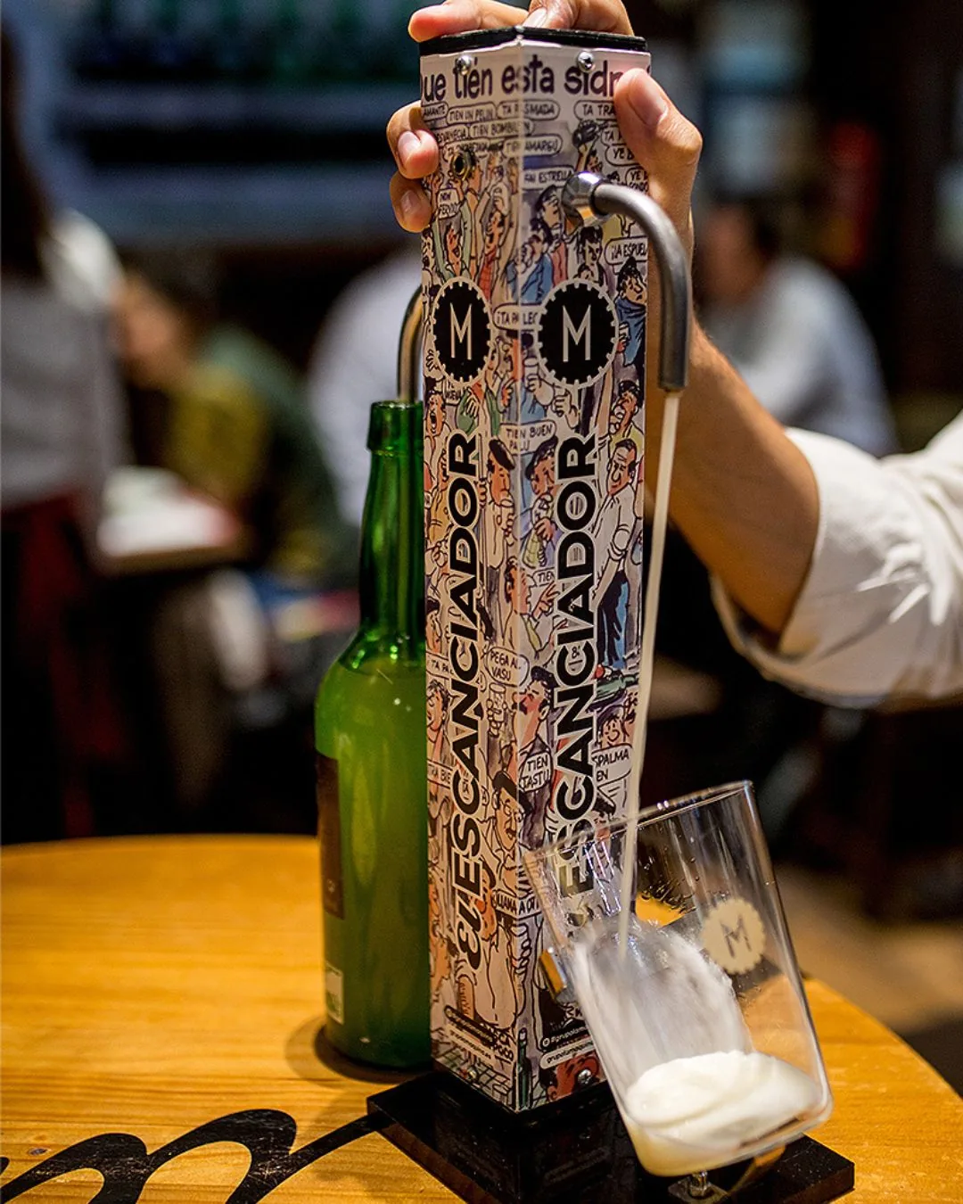 Asturian cider poured from a mechanical escanciador into a glass, beside a green cider bottle