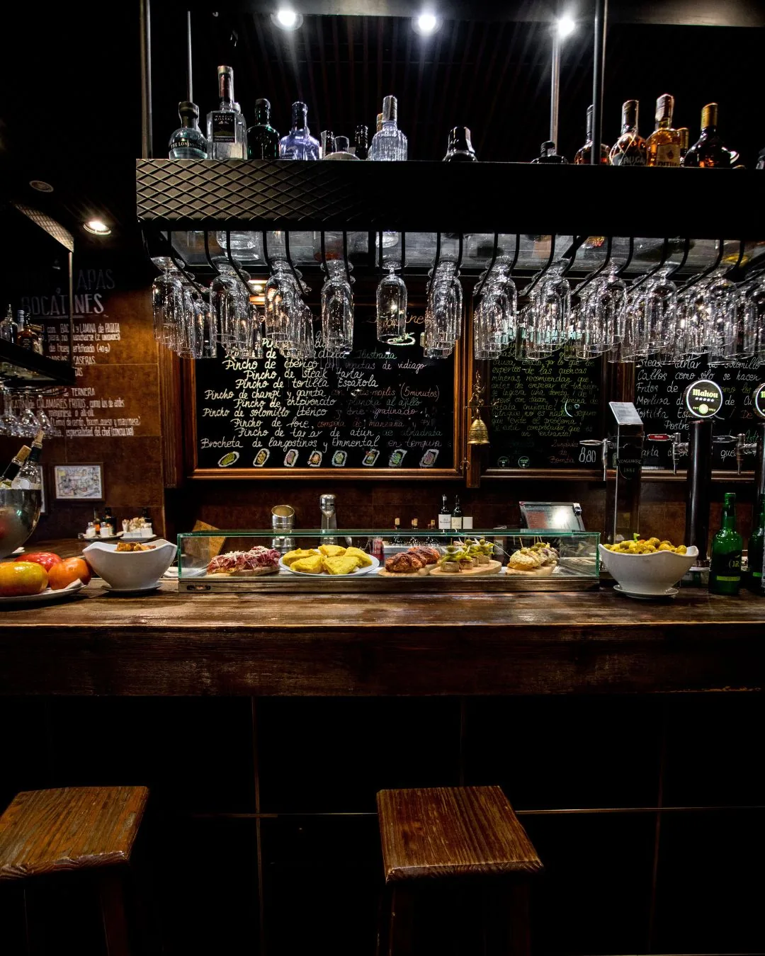 The bar counter with a glass case of pinchos, hanging glasses and a chalkboard menu