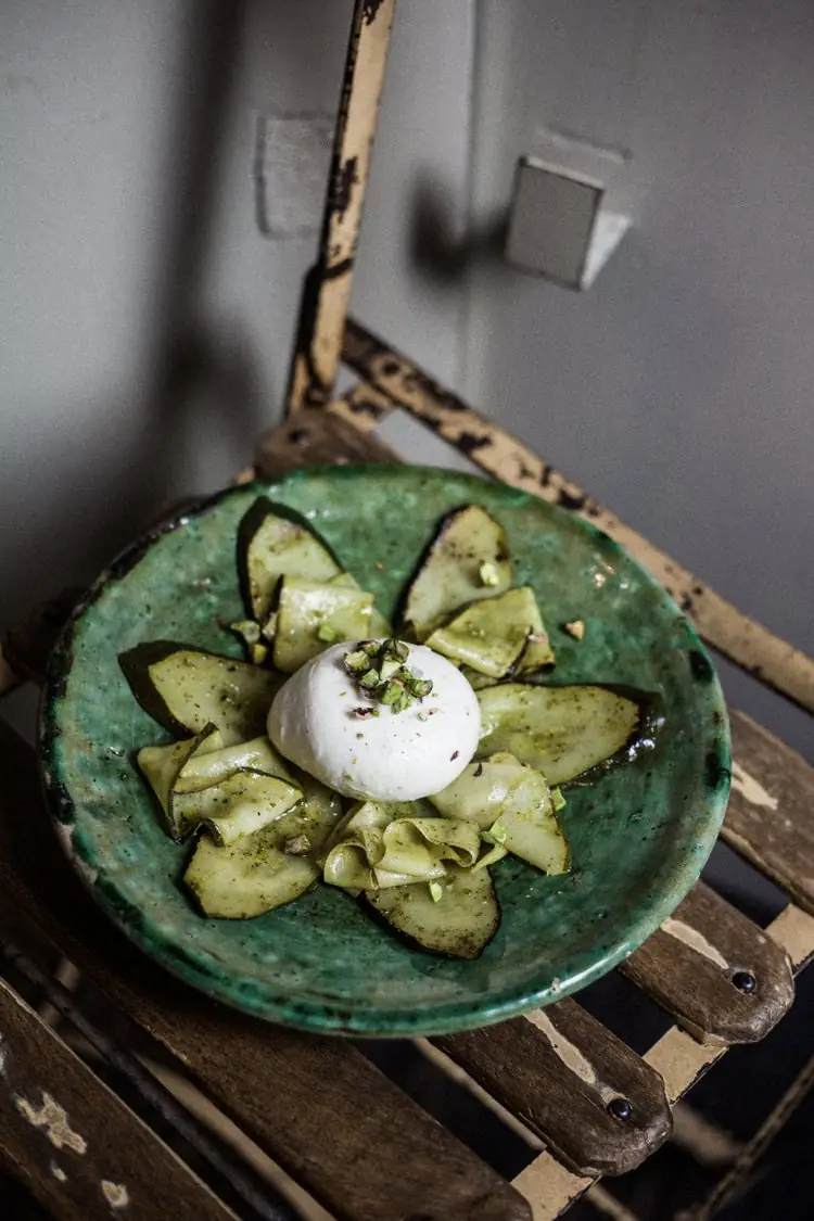 Burrata topped with chopped pistachios on a bed of thin pear and green herb-oil slices, arranged in a flower shape on a green ceramic plate resting on a rustic metal chair