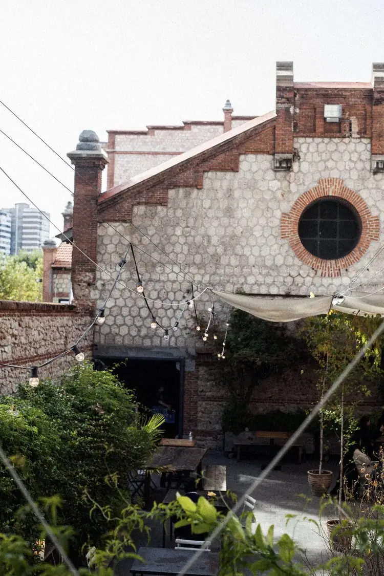 Exterior terrace of the venue with a brick-and-stone industrial building, a round brick-framed window, string lights and wooden benches among greenery