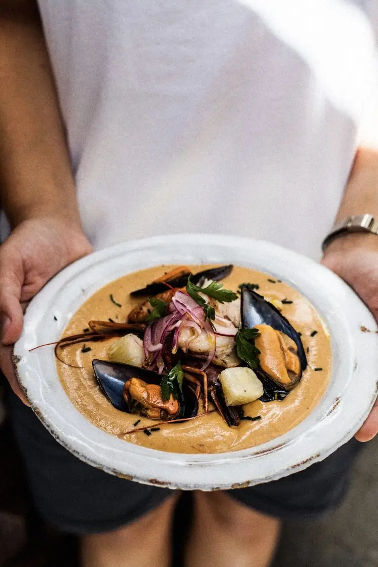 Deep white plate of mussels, prawns and fish in a creamy orange seafood broth topped with pickled red onion and parsley, held by a person in a white shirt