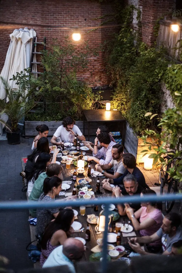Diners seated along a long wooden table on the venue's brick-walled terrace at dusk, with candles, beer bottles and string lights