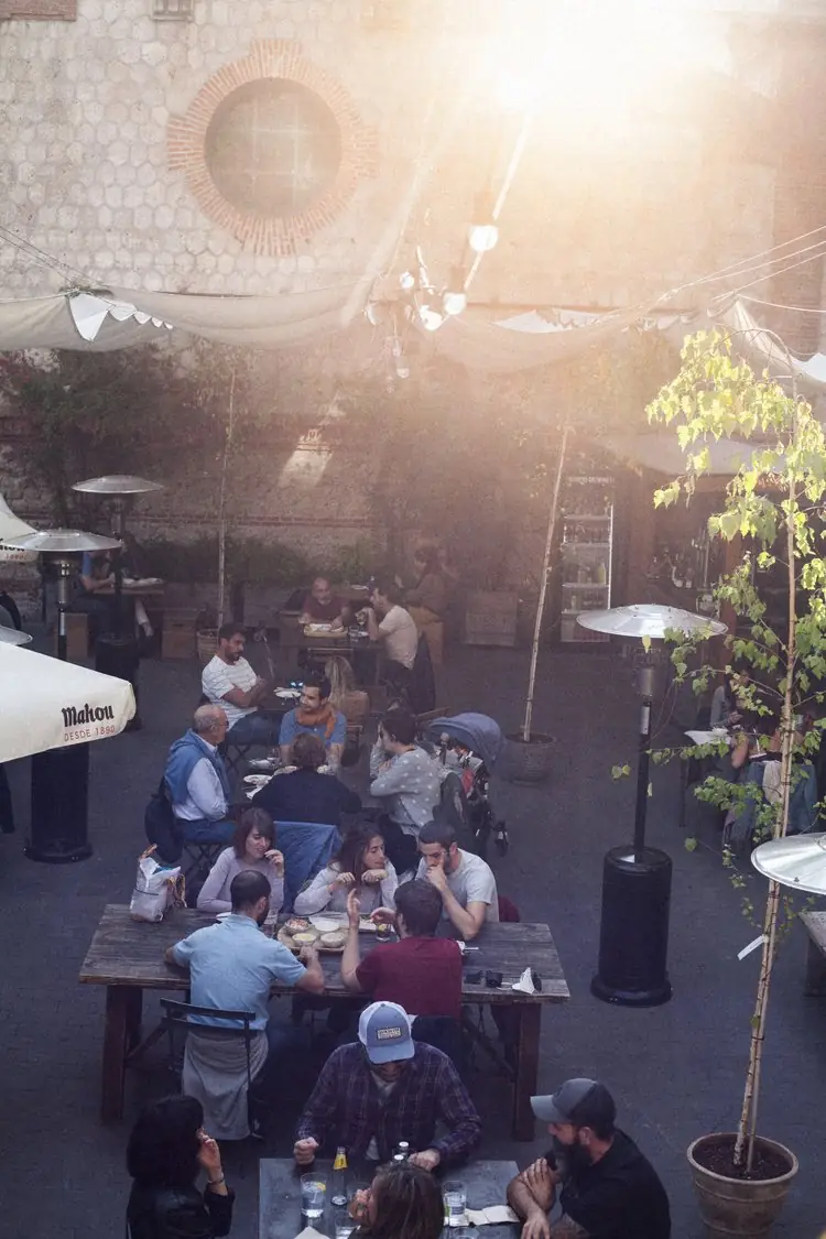 Patrons at wooden tables in the venue's courtyard terrace with patio heaters, umbrellas and hanging canvas shades in warm afternoon sun