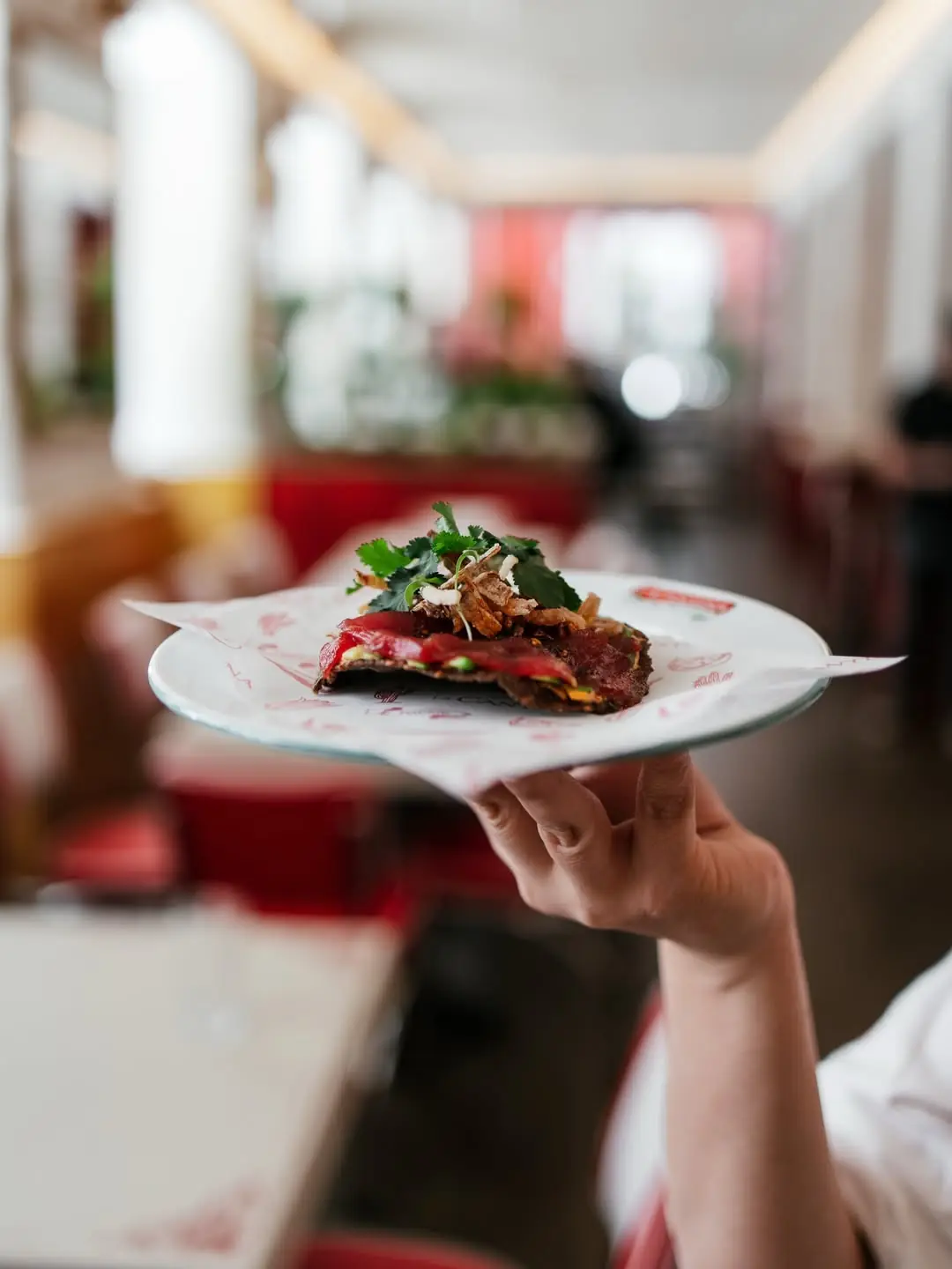 A hand holding a plate with a tostada topped with sliced meat, herbs, and crispy garnish.