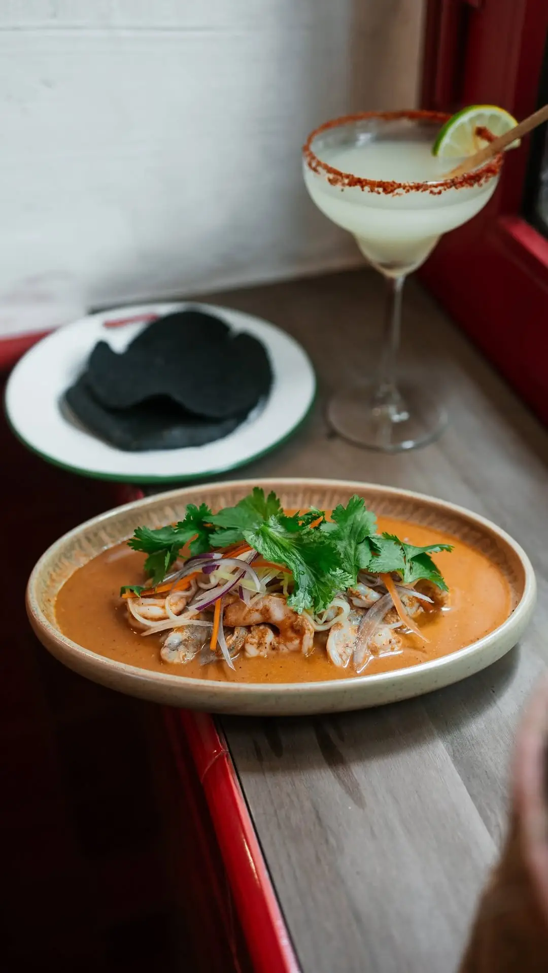 A shrimp aguachile dish topped with cilantro and vegetables, with a margarita and blue tortillas in the background.