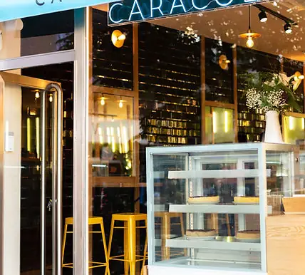 Cafe storefront with glass windows, yellow bar stools outside and a pastry display case, signage reading CARACOL visible above
