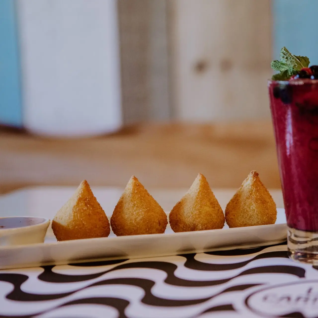 Four golden fried coxinha croquettes on a white plate with dipping sauce, next to a berry cocktail garnished with mint, La Carioca