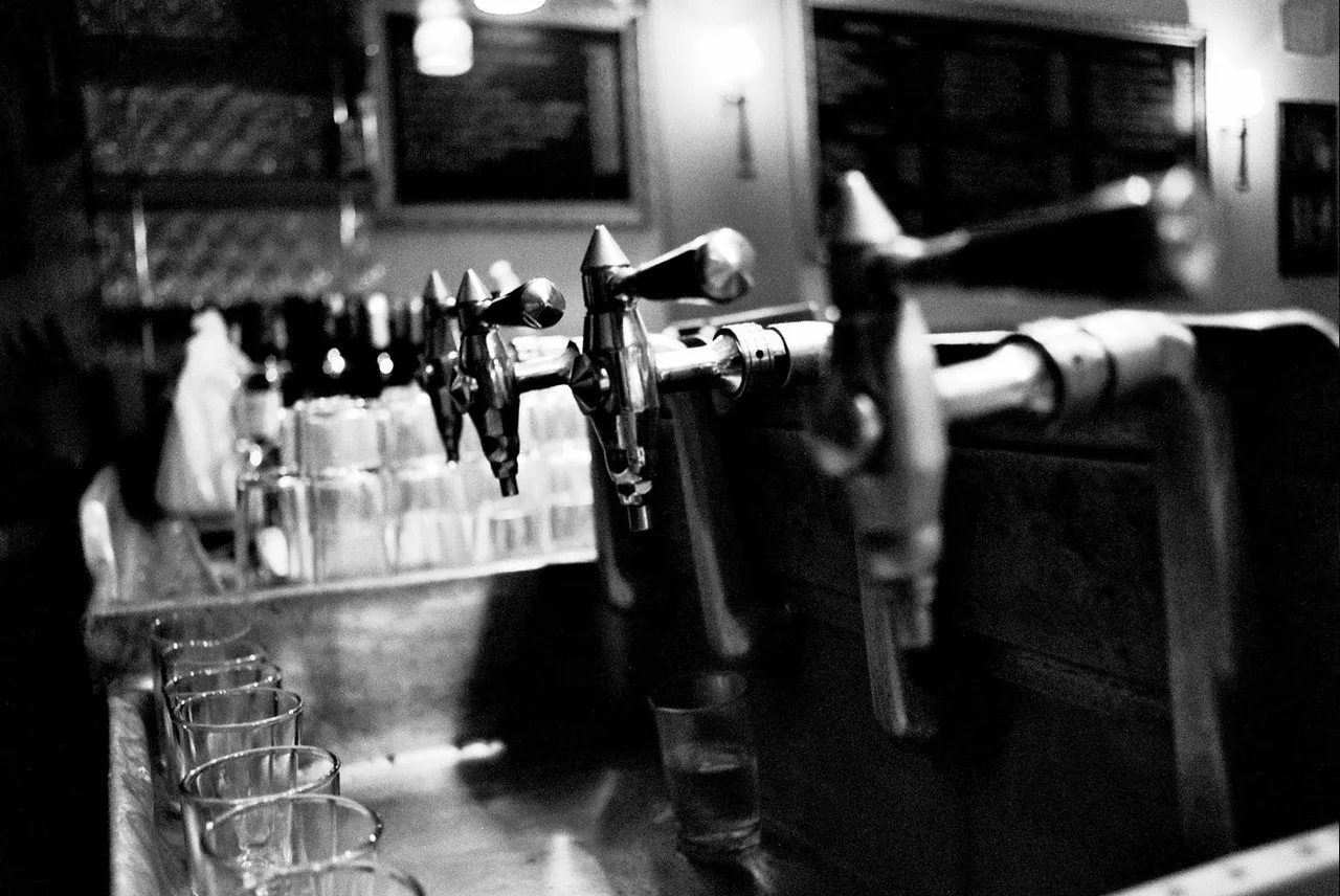 Black-and-white view along a bar counter showing chrome beer taps, rows of upturned glasses and a bar towel, with framed pictures on the wall behind