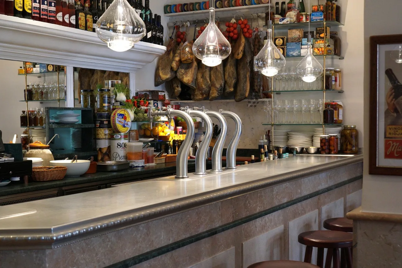 Long marble bar counter with four curved chrome beer taps, whole cured hams and strings of tomatoes hanging behind, shelves of tinned conservas and stacked glassware, and stools along the front