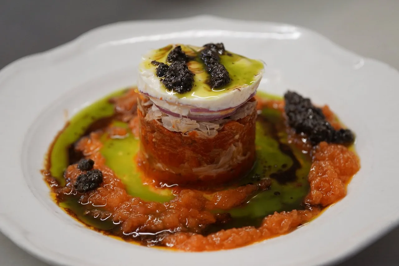A plated timbale of crushed tomato and shredded tuna with red onion, topped with a round of soft goat cheese, black olive tapenade and green herb oil on a white plate