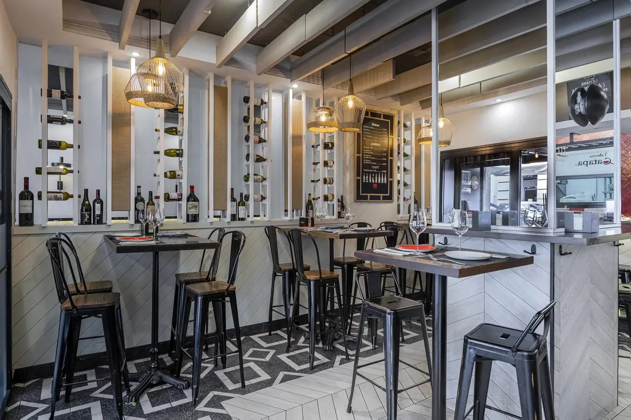 A narrow tavern room of high wooden tables and black metal stools set with red placemats and wine glasses, under a wall of vertical white wine racks and a chalked beer list