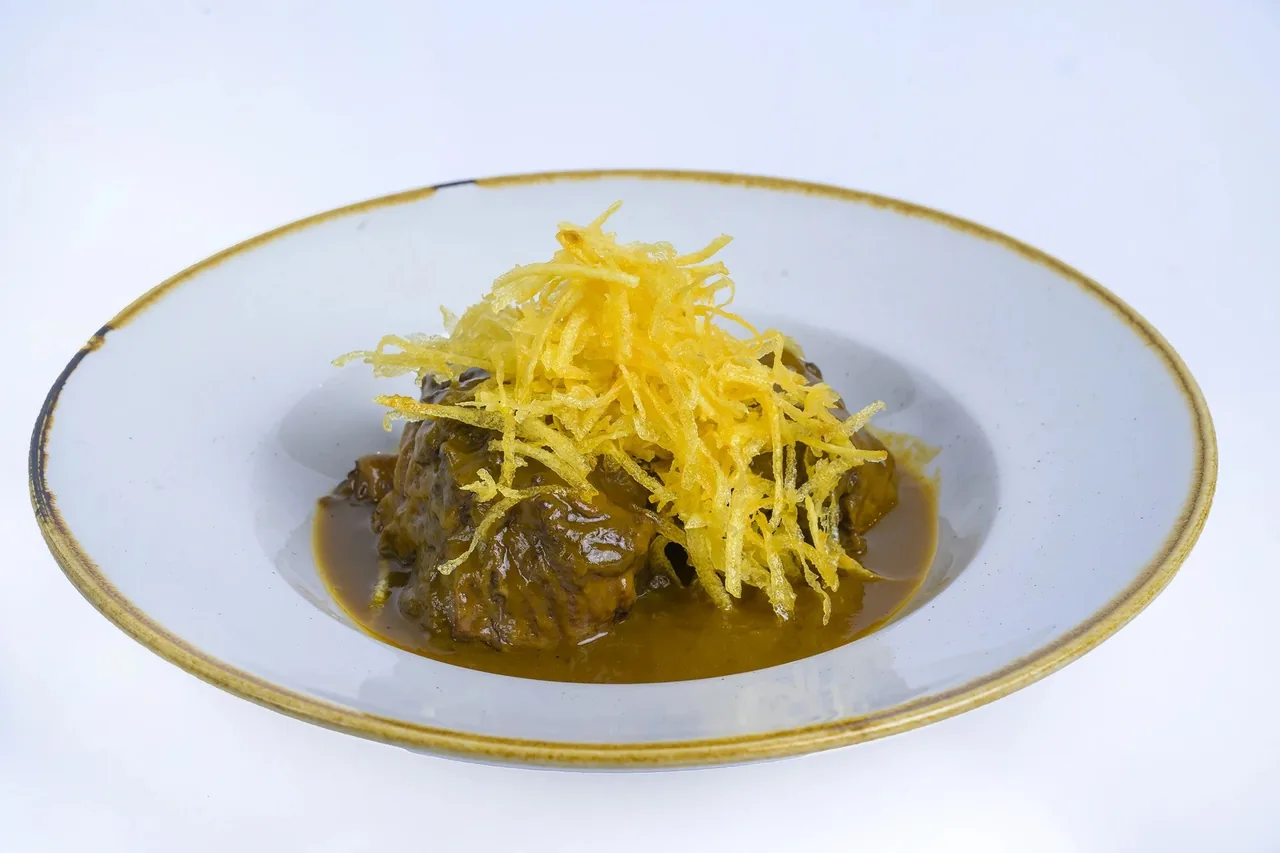 A shallow white bowl of braised meat in dark sauce topped with fine potato strips