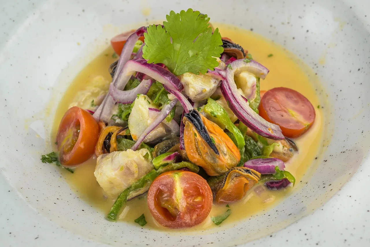 A shallow bowl of salad with red onion, tomato, mussels and green leaves in a yellow dressing