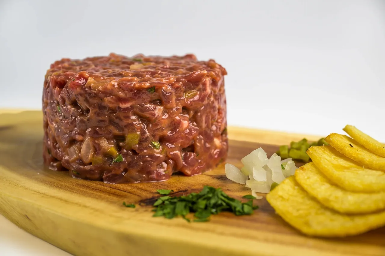 A round mound of minced red meat on a wooden board beside fried slices