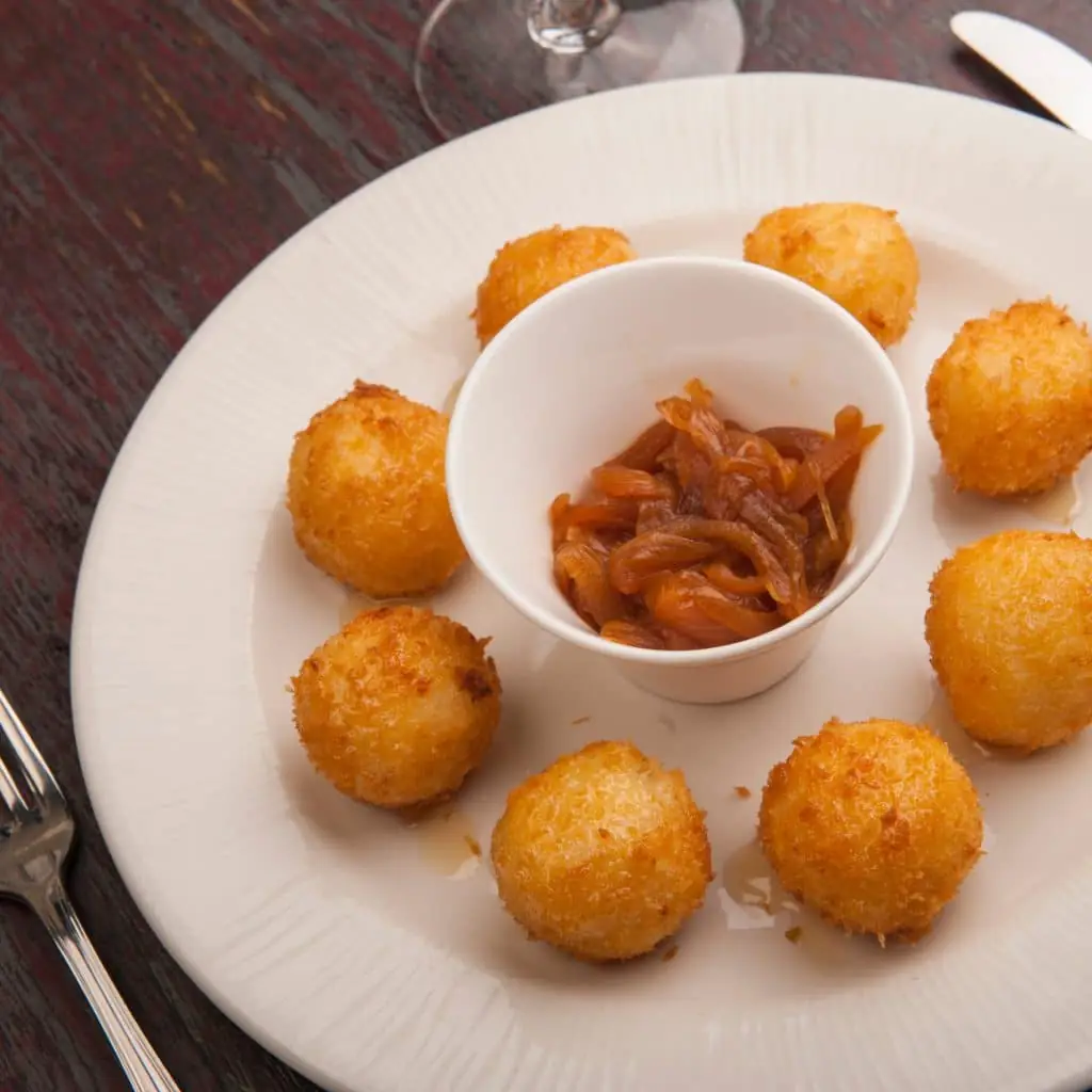 Eight golden fried cheese balls arranged around a white pot of caramelized onions on a white plate