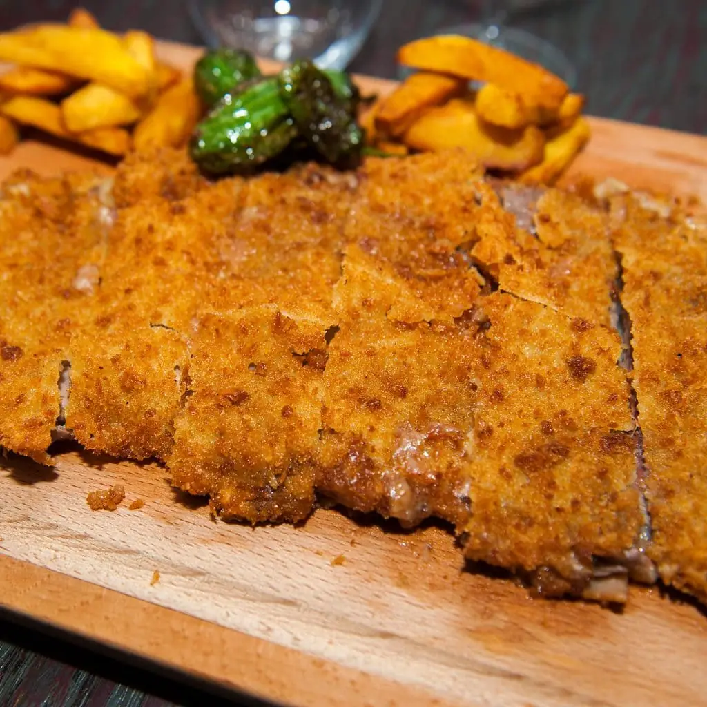 Sliced breaded veal cachopo/cutlet on a wooden board with thick-cut fries and blistered padron peppers