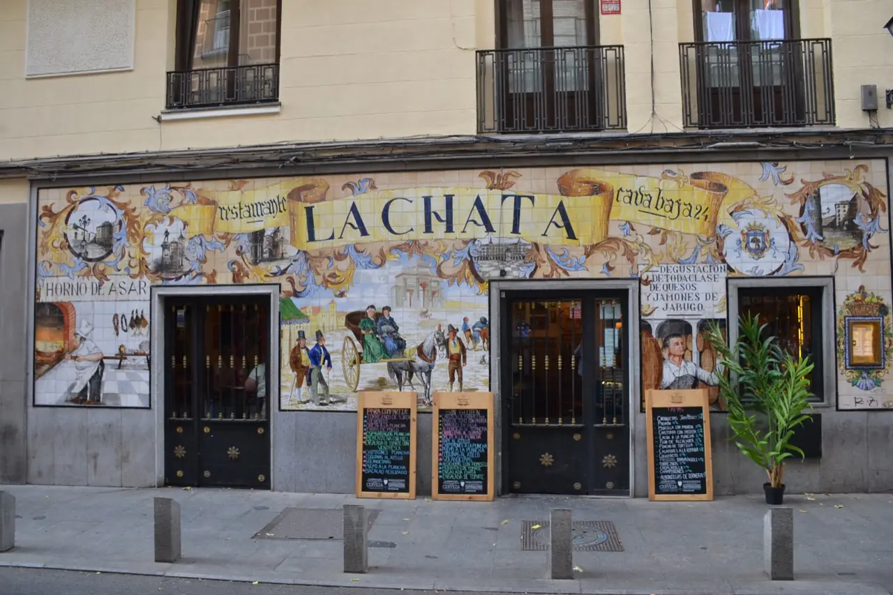 Daytime exterior of Restaurante La Chata at Cava Baja 24, showing its ornate painted ceramic tile facade with the restaurant name and scenes of old Madrid, chalkboard menus, and a potted plant by the entrance.