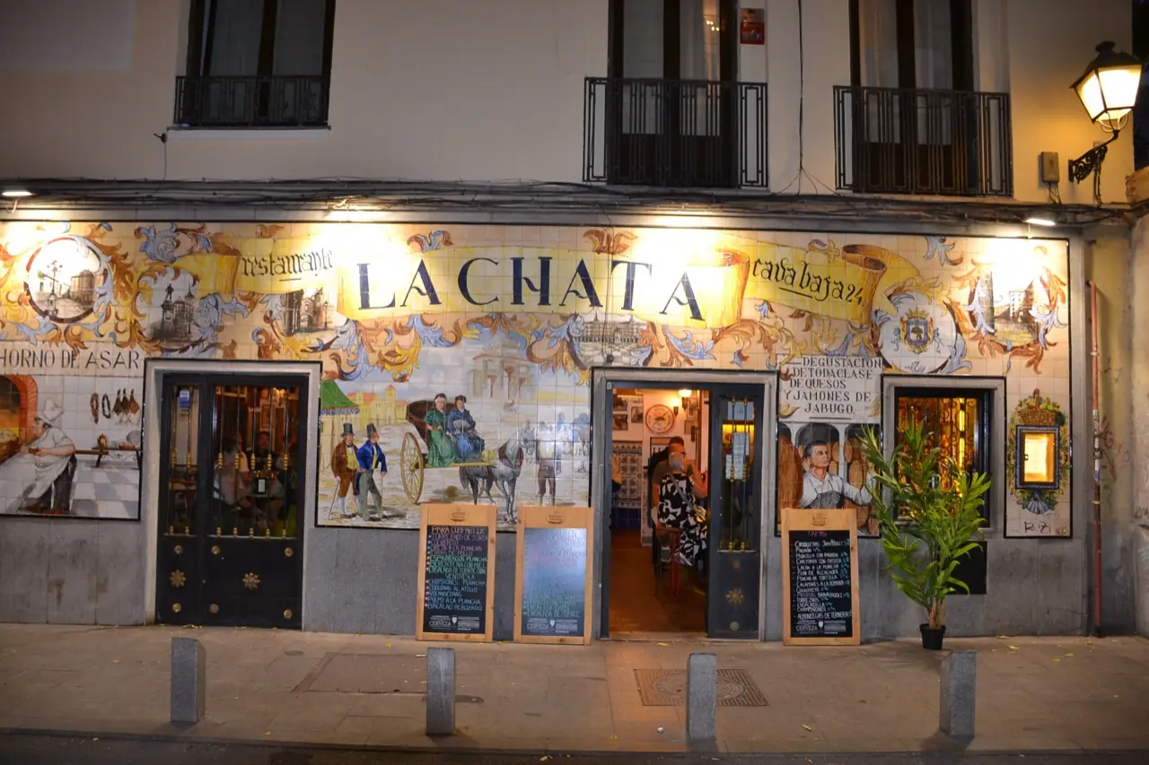 Nighttime exterior of La Chata's illuminated tiled facade with diners visible seated inside through the open door.