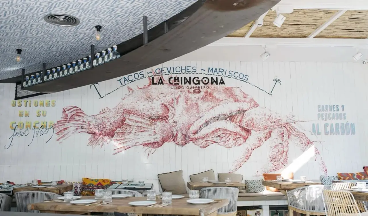 Interior dining room of La Chingona with a large wall mural of a crab and text reading Tacos, Ceviches, Mariscos, La Chingona Estado Guerrero, above set wooden tables with woven chairs and patterned cushions.