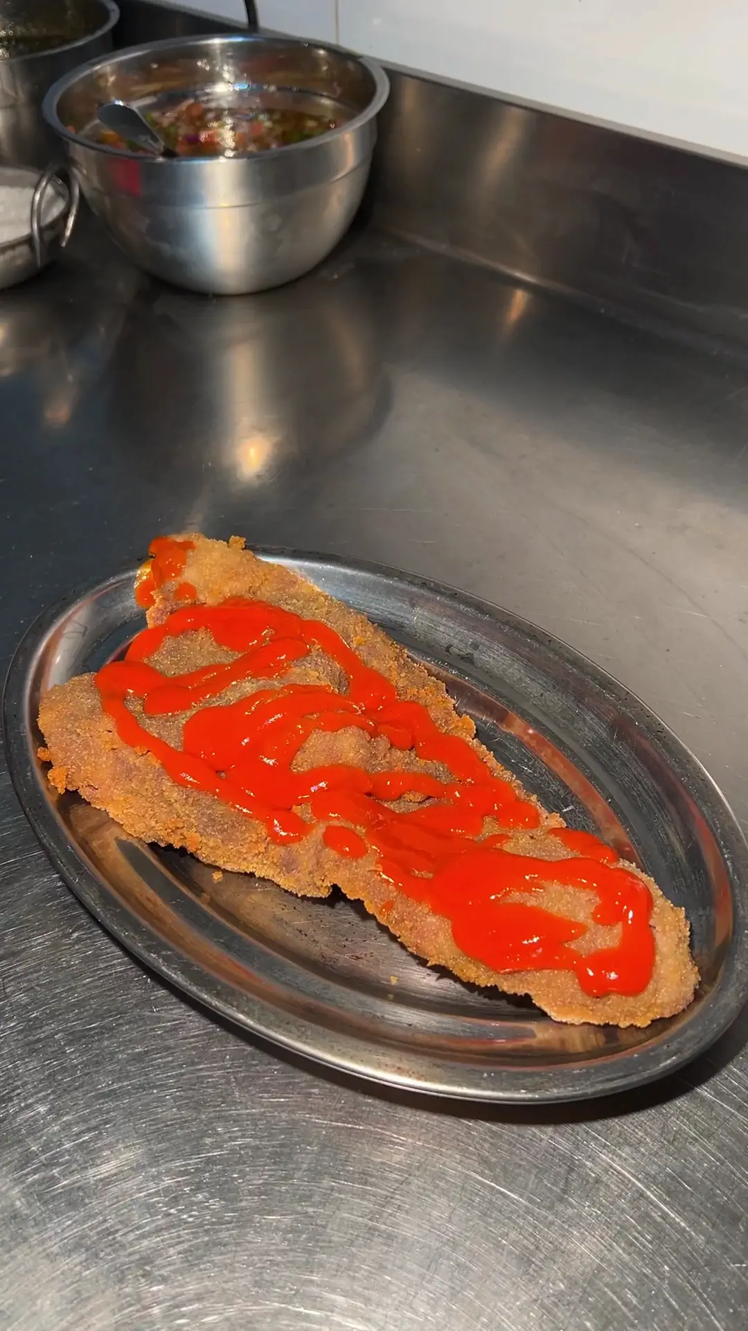 Breaded milanesa with a zigzag of red sauce on a metal tray.