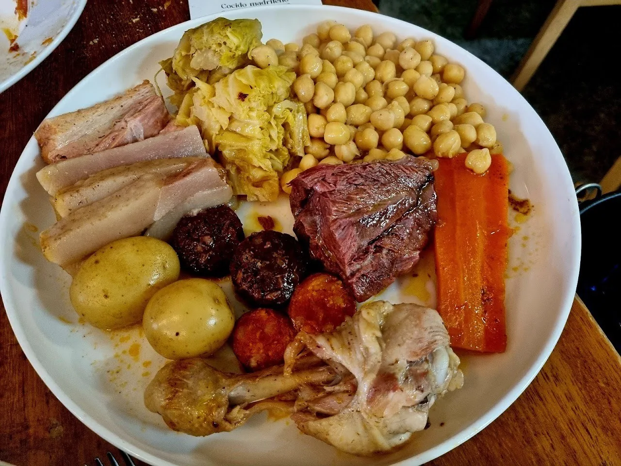 A white plate of chickpeas, cabbage, potatoes, carrot, boiled pork belly, black pudding and chorizo