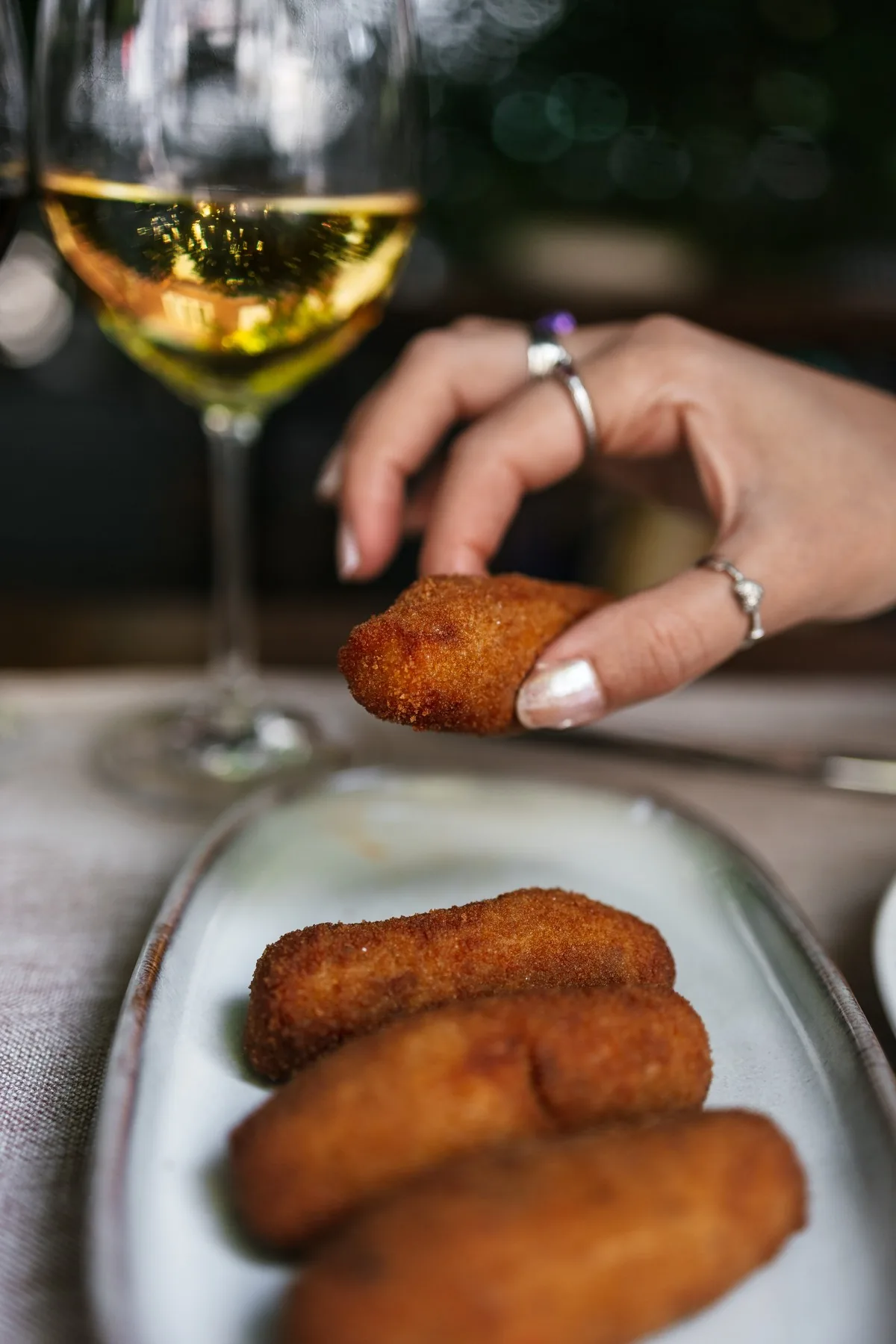 A hand reaching for fried croquettes on a plate with a glass of white wine