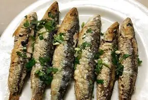 Six grilled sardines topped with chopped parsley and garlic, arranged in a row on a white plate at La Cova Fumada.