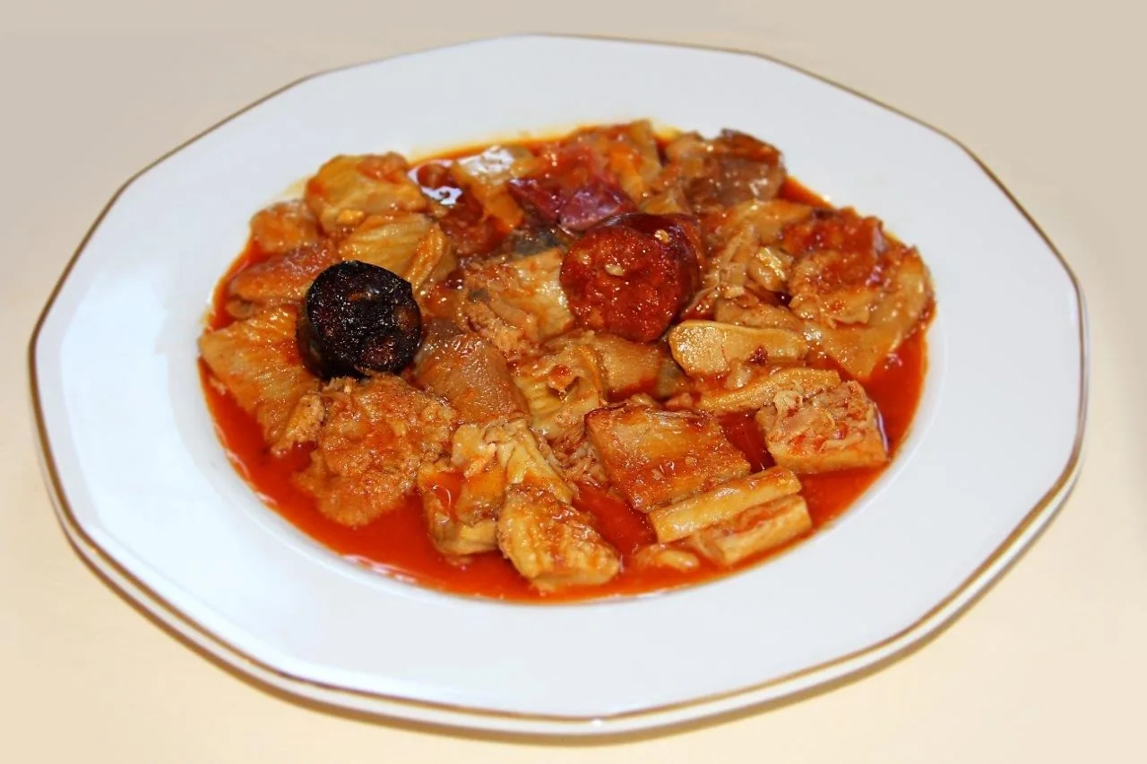 A white plate of tripe stew in red sauce with slices of chorizo and black pudding