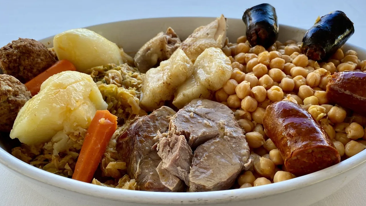 A white bowl filled with chickpeas, boiled beef, potatoes, cabbage, carrot, chorizo and black pudding