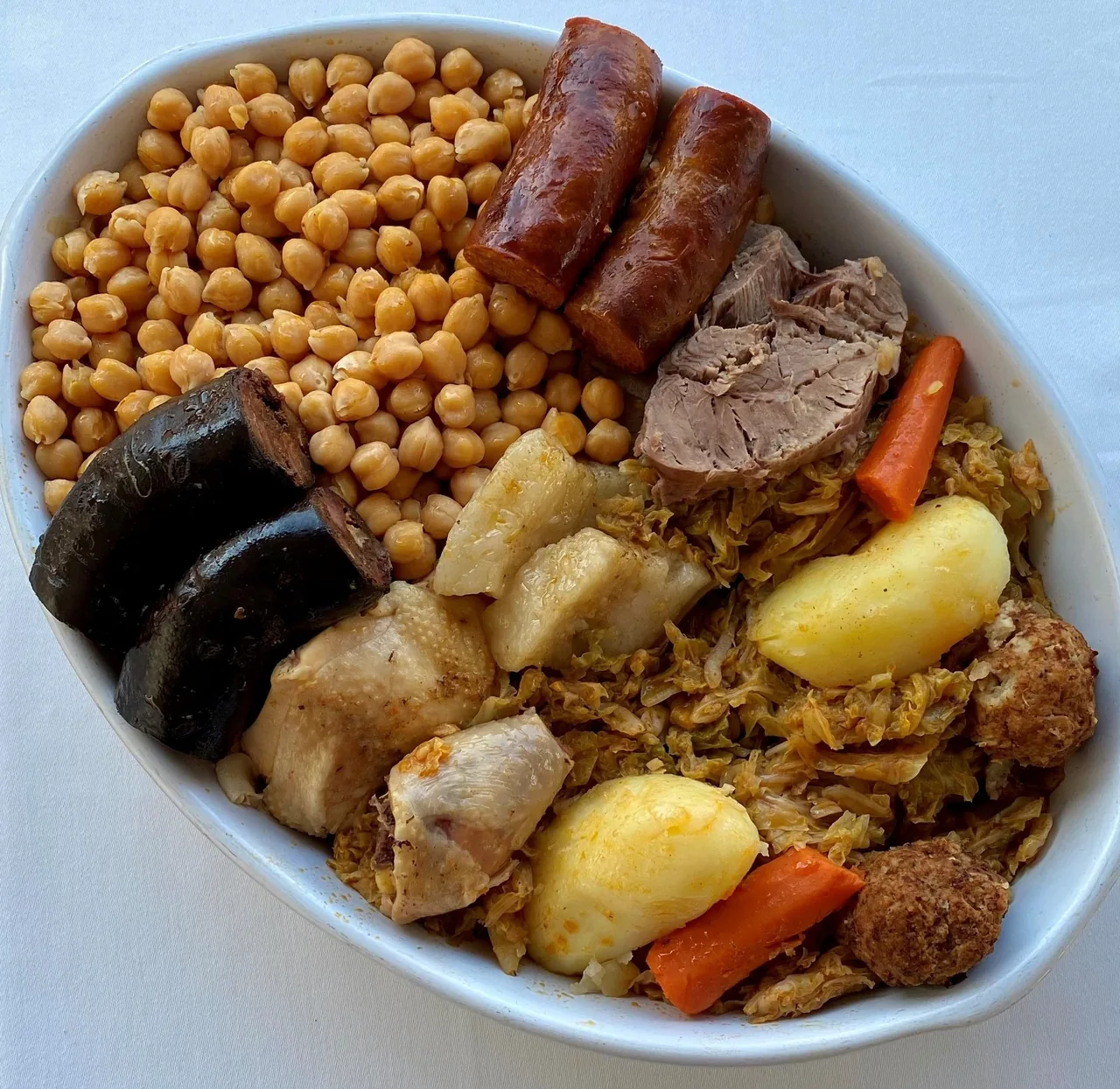 A close view of a white oval bowl of chickpeas with chorizo, black pudding, boiled meats, cabbage, potato and carrot