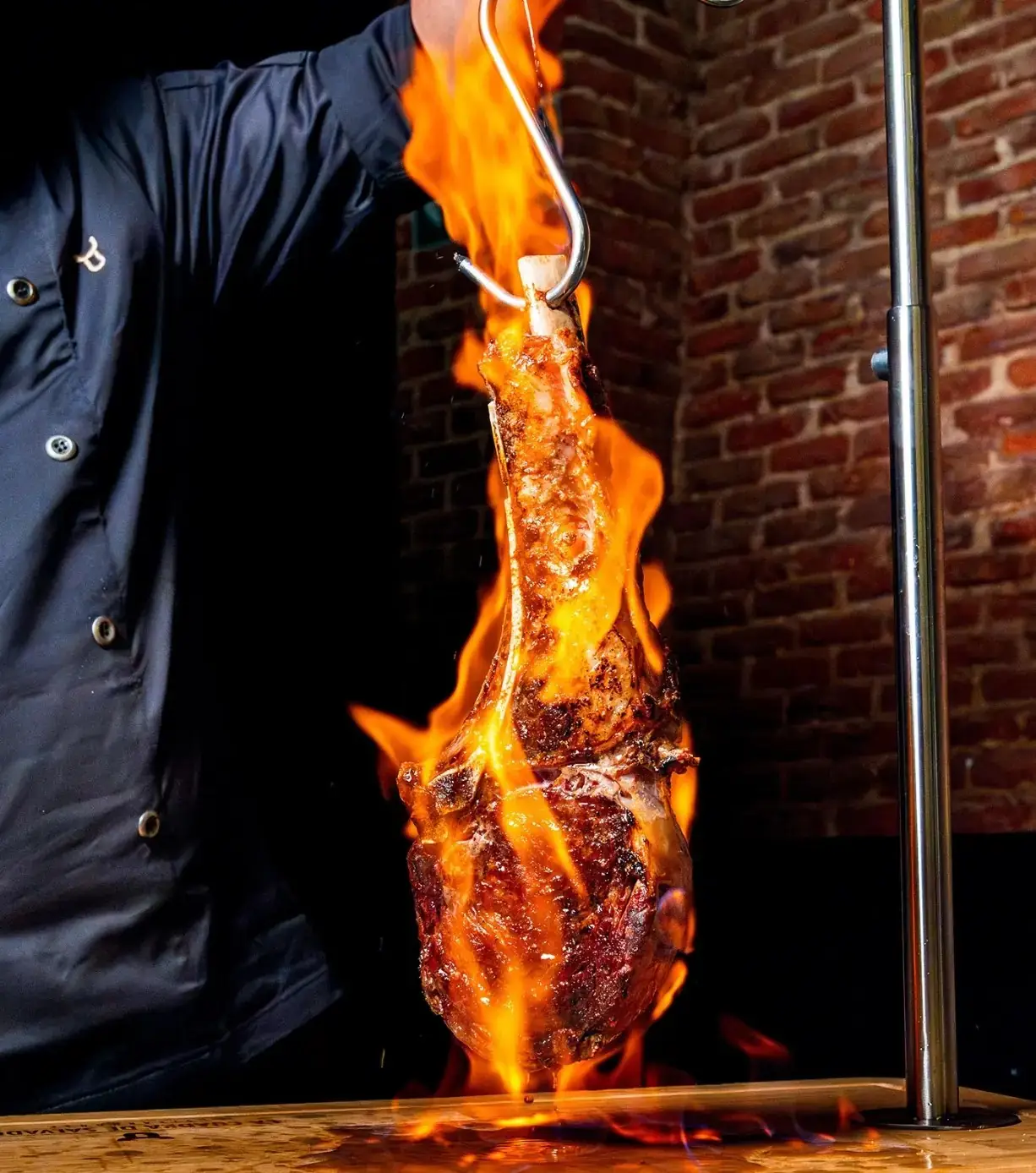 A charred tomahawk steak hanging from a hook engulfed in flames over a wooden board against a brick wall