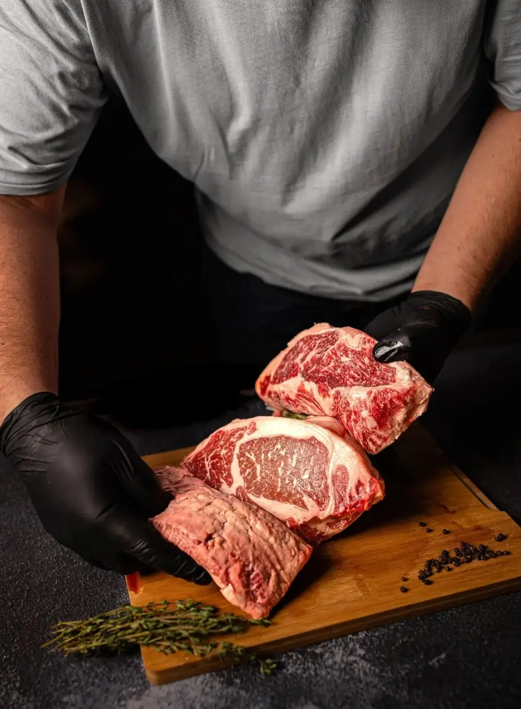 Gloved hands presenting three thick marbled raw beef cuts on a wooden board with thyme and peppercorns