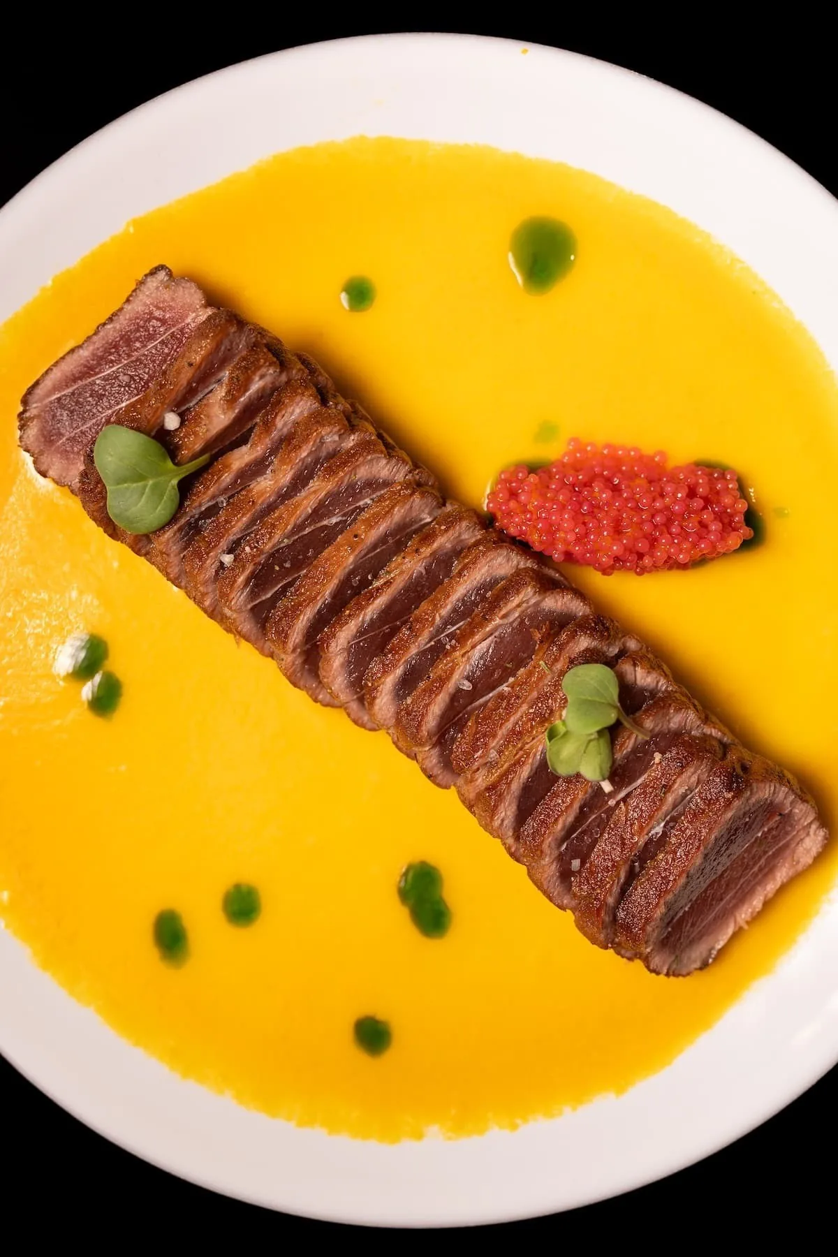 Thin slices of grilled beef on a plate of bright yellow sauce with green dots and a red garnish