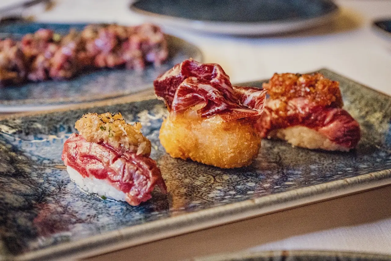 Three small plated bites of seared meat topped with crispy garnishes on a slate tray