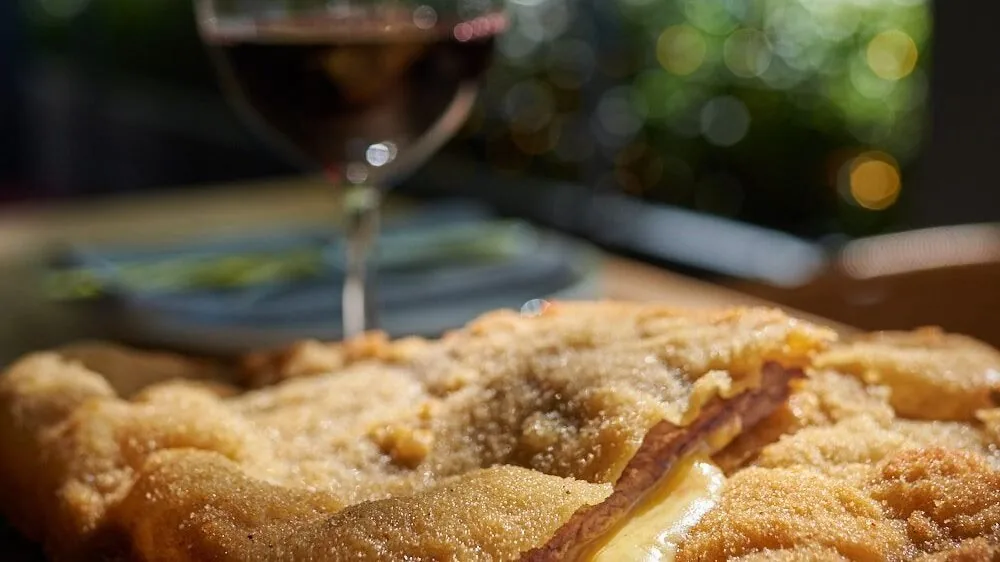 Close-up of a breaded cutlet oozing melted cheese, with a glass of red wine and a plated side blurred behind