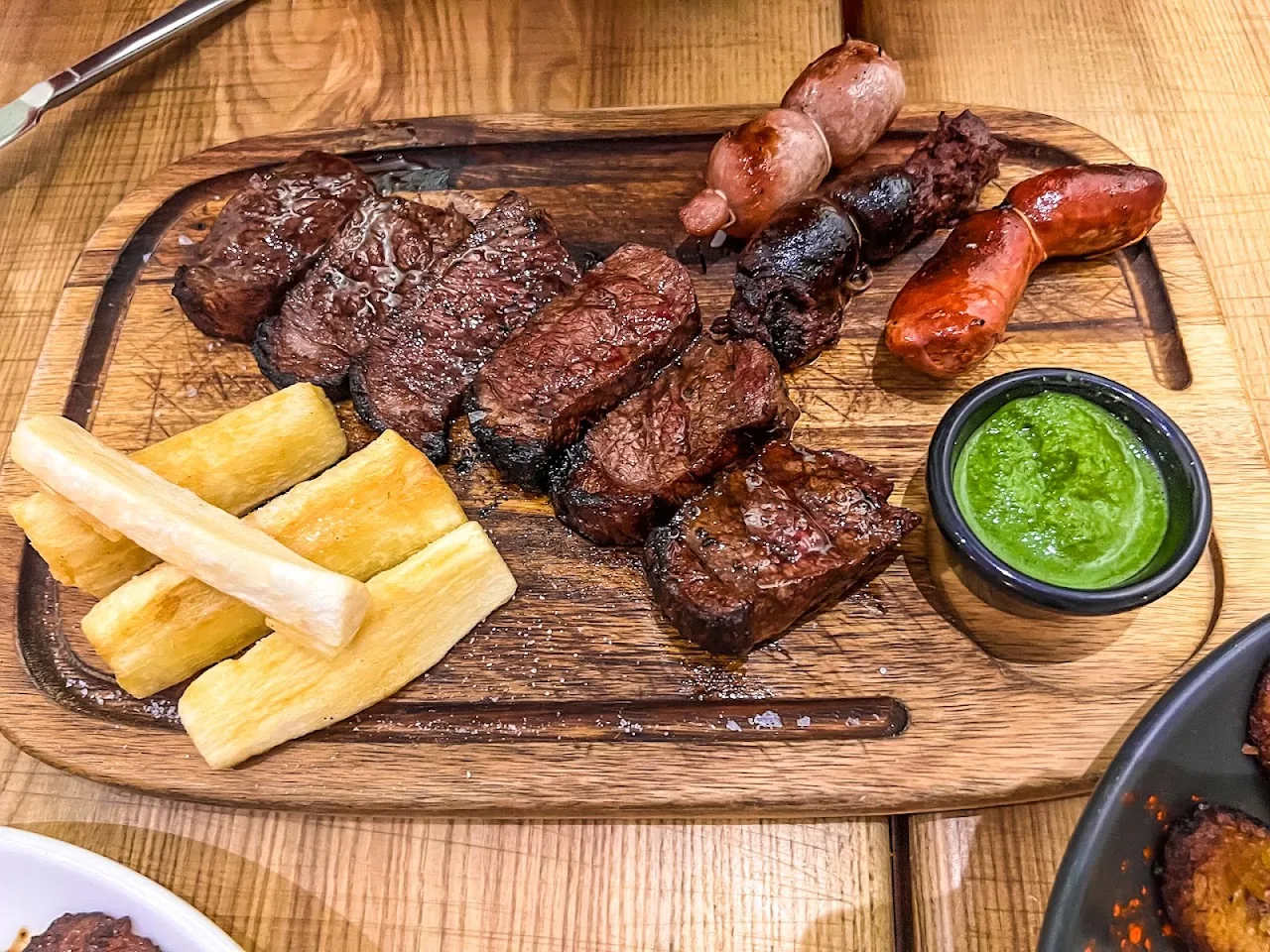 A wooden board of grilled meats, sausages, and fried yuca with green sauce
