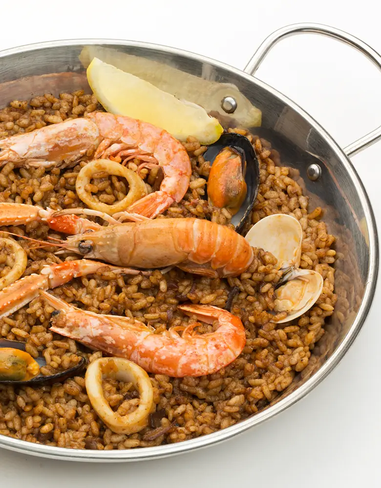 Seafood paella in a steel pan with shrimp, mussels, clams, and calamari rings, topped with a lemon wedge, at La Cuina de Laietana