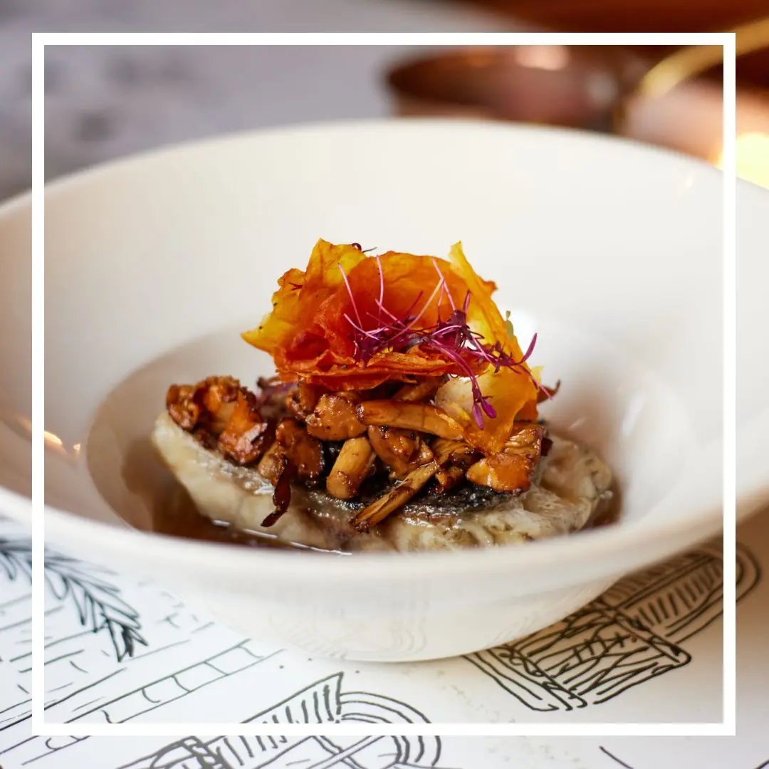 Pan-seared fish fillet topped with sautéed wild mushrooms and a crisp orange garnish in a white bowl, at La Dama