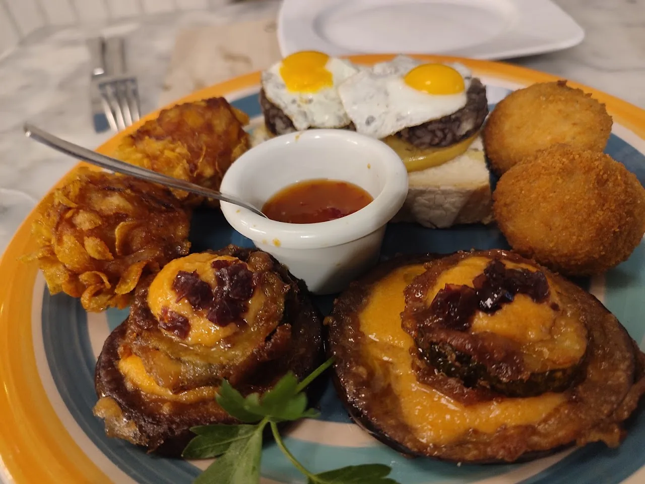 A painted plate of tapas: croquettes, fried eggs on toast, a crisp fried bite and a dish of red sauce