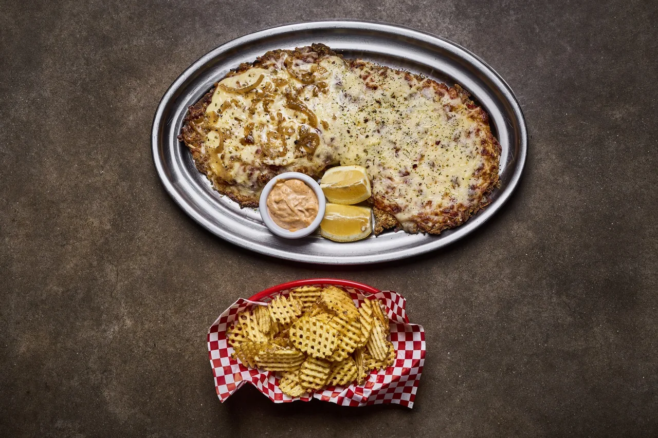 A cheese-topped breaded cutlet on a metal tray next to a basket of waffle fries