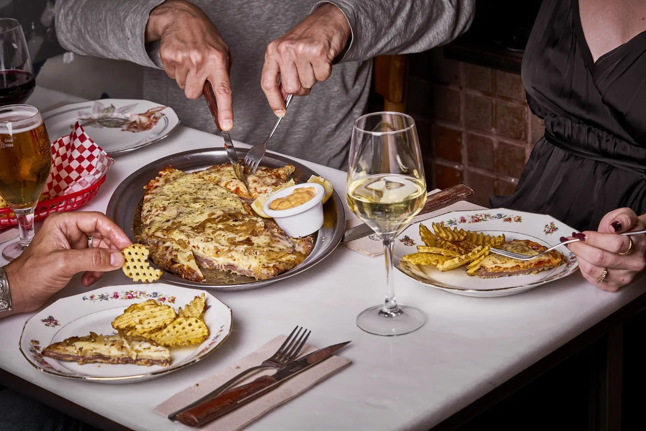 A cheese-topped breaded cutlet being cut at a table with plates of waffle fries